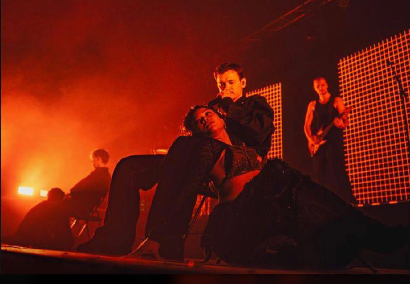 A theatrical moment during a slow song, one singing seated while a dancer reclines across his lap on a stage washed in red and orange haze
