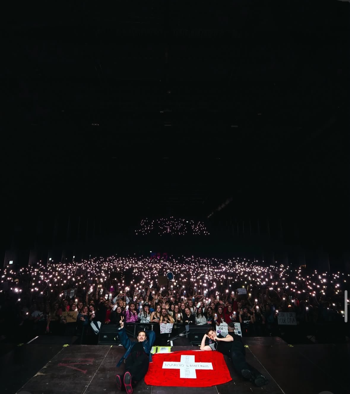 Marcus & Martinus lying at the edge of the stage with a Swiss flag made by fans, the crowd holding phone lights behind
