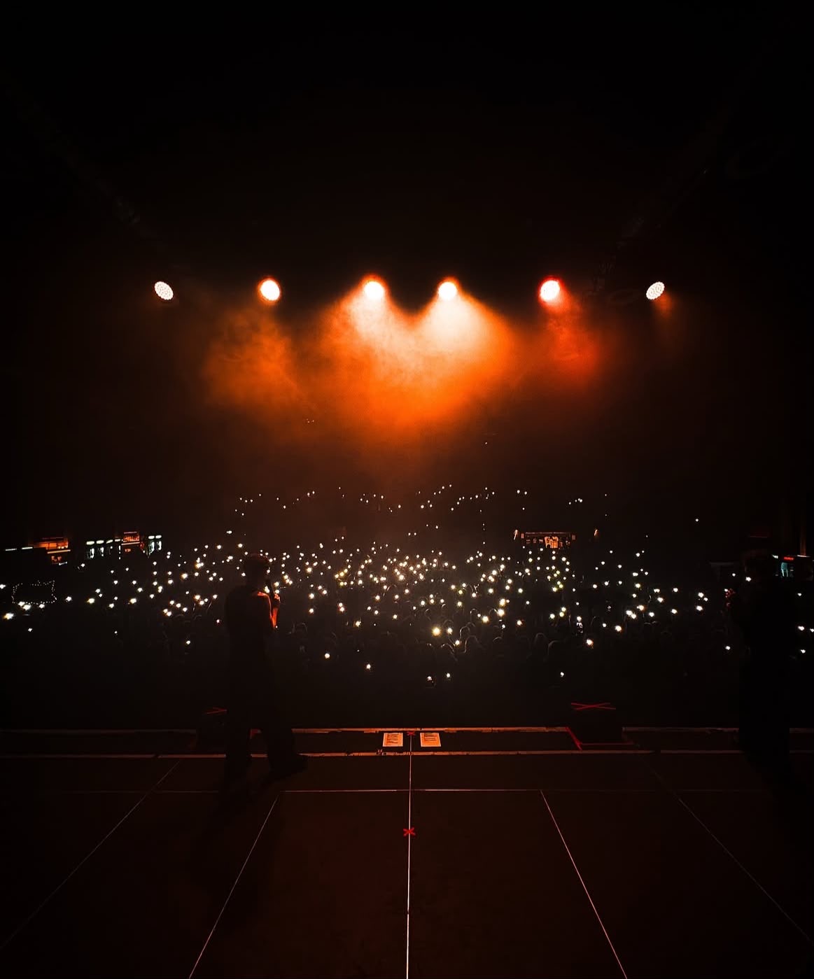 A performer silhouetted on stage facing an arena full of phone lights