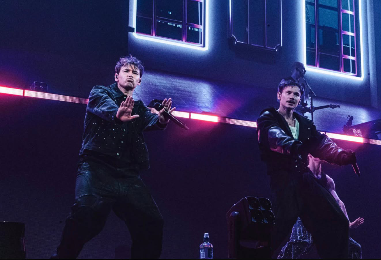 Marcus & Martinus mid-choreography in dark jackets in front of neon-outlined windows glowing pink and cyan