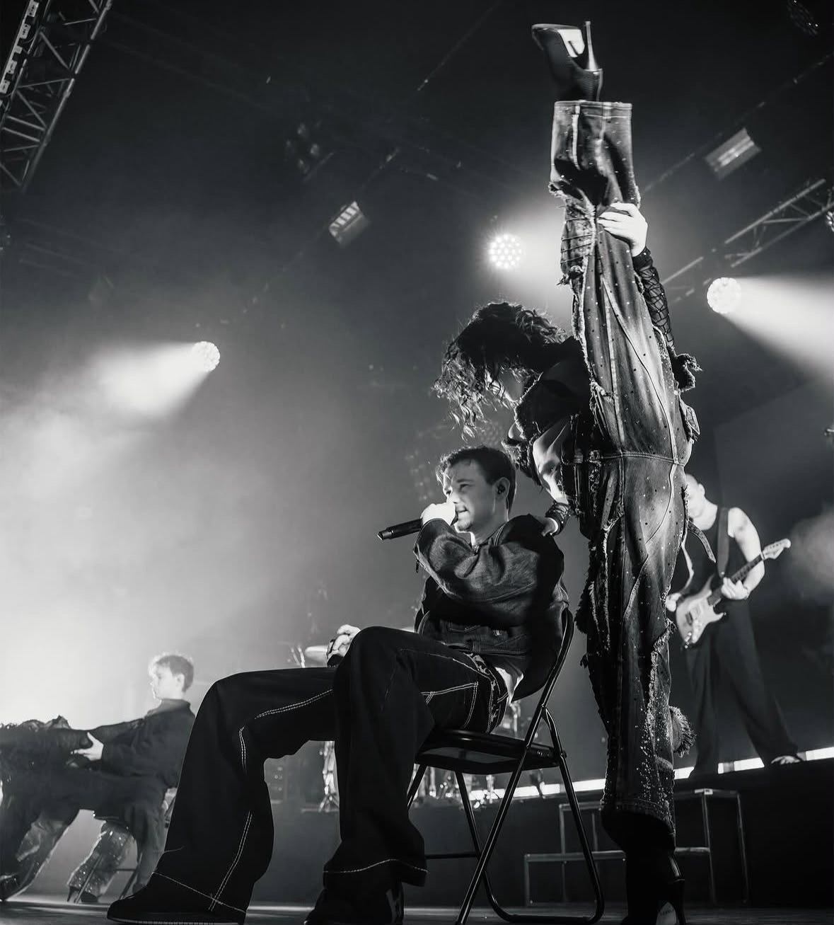 Singing seated on a chair while a dancer holds a vertical split behind, in black and white