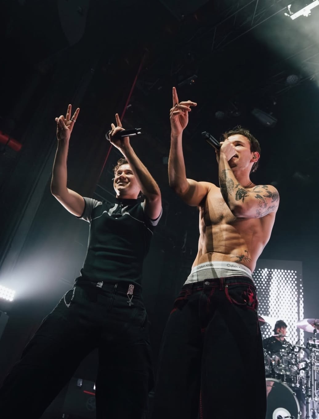 Marcus & Martinus hyping the crowd from a low angle on stage, microphones in hand and signs raised against a glowing white LED backdrop